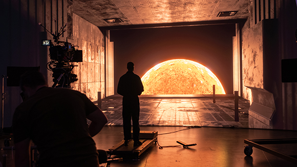 A silhouette of a person stands before a massive, glowing orange sun on a dark film studio set.