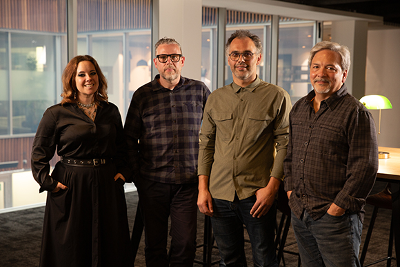 Four adults standing in a row indoors, smiling at the camera.