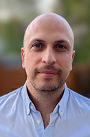 A bald man with blue eyes and light stubble wearing a light blue button-down shirt.