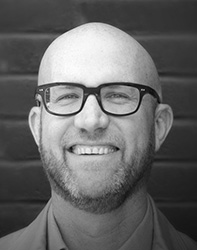 Black and white headshot of a bald man with glasses and a beard, smiling.