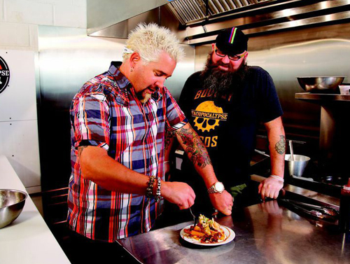 A man with spiky blonde hair in a plaid shirt plates food in a professional kitchen.