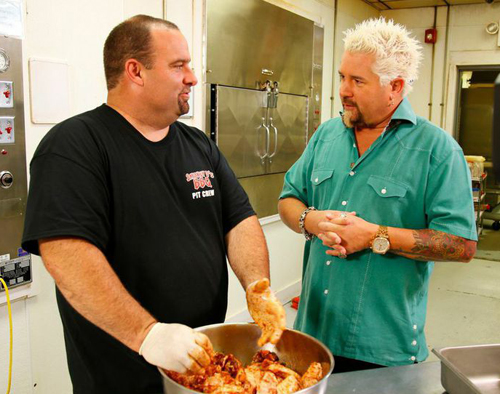 A man in a black t-shirt holds a bowl of food while talking to a man in a teal shirt in a kitchen.