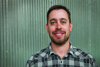 A smiling man with short brown hair and a beard wearing a grey plaid shirt.