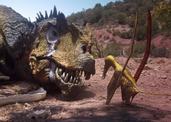 A large Tyrannosaurus Rex model faces a small Pterosaur model in a rocky, hilly terrain.