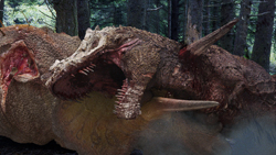 A large, brown, scaly creature with an open mouth and sharp teeth lies in a forest.