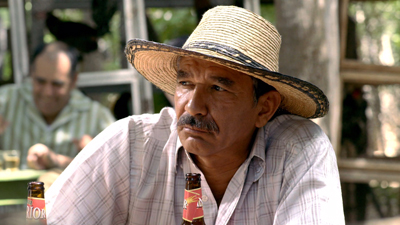 A man with a mustache wearing a wide-brimmed straw hat sits at an outdoor table with beer bottles.