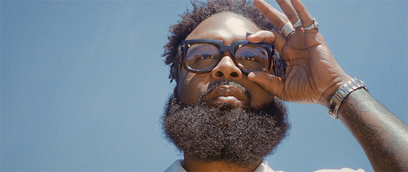 A man with a thick beard and glasses looks down while adjusting his frames against a clear blue sky.