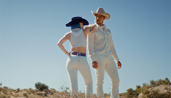 A person in a blue hat and white crop top stands next to a person in a white cowboy hat and white button-down shirt.