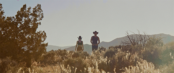 Two people in hats stand in a brushy desert landscape under a clear sky.