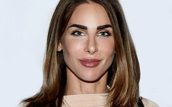 A woman with long brown hair and light eyes looks directly at the camera against a white background.