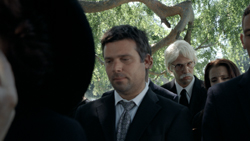 A man in a dark suit and patterned tie looks down while others stand behind him in a wooded area.