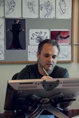 A man sits at a desk looking at a computer monitor with sketches pinned to a board behind him.