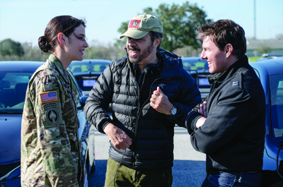 A woman in military camouflage, a man in a puffer jacket and cap, and a man in a black jacket talk together.