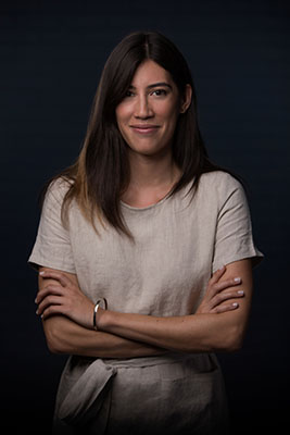 A woman with long dark hair and a beige linen top stands with her arms crossed against a dark background.