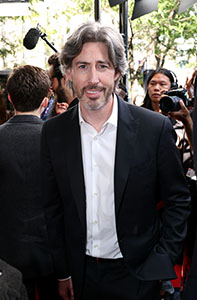 A man with wavy grey hair wearing a black suit and white shirt smiles at an outdoor event.