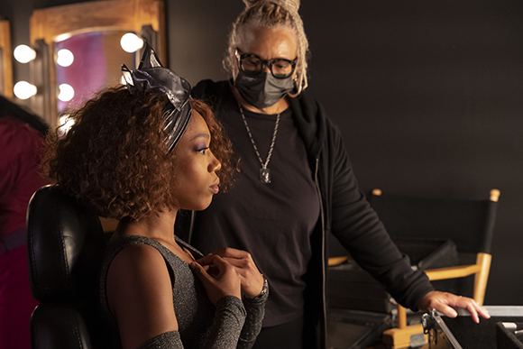 A person in a black mask and glasses adjusts the makeup of a woman with curly hair sitting in a chair.