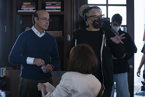 A man in a blue sweater speaks to a woman wearing a black mask and headphones in a room with bookshelves.