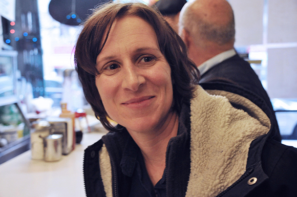 A woman with short brown hair smiles while wearing a dark jacket with a fleece lining.