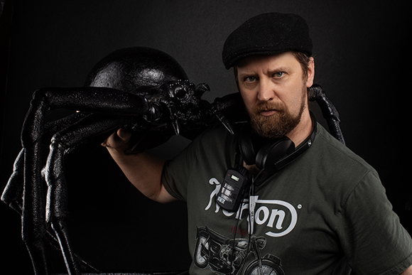A bearded man in a black cap and green t-shirt poses behind a large, black, textured spider sculpture.