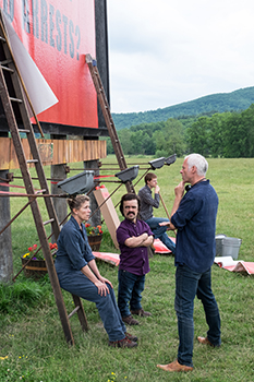 Four people stand in a grassy field near a large red billboard and wooden scaffolding.