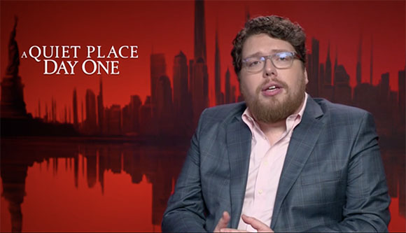 A man with glasses and a beard wearing a suit jacket speaks in front of a red city skyline background.