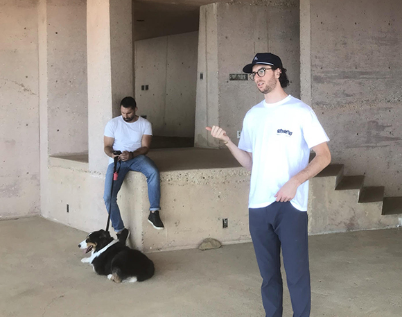 A man in a white t-shirt and cap stands in the foreground while another man sits with a dog in the background.