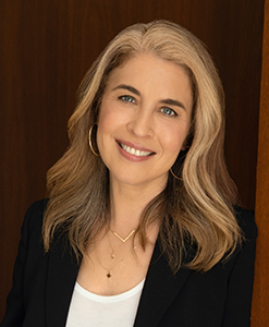 A smiling woman with shoulder-length blonde hair, wearing a black blazer and gold hoop earrings.