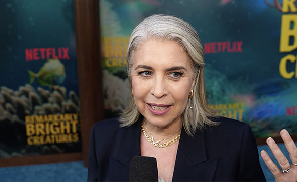 A woman with shoulder-length grey hair and gold jewelry speaks during an interview.