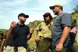 Three men in casual clothing standing together outdoors near a hillside.