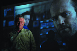 A man smiles in a dark room in front of a large cinematic close-up of a man's face.