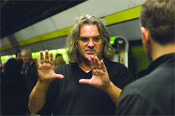 A man with long wavy hair and glasses gestures with both hands raised in front of him.
