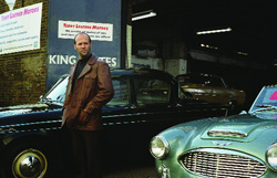A man in a brown jacket stands beside a black vintage car in a garage setting.