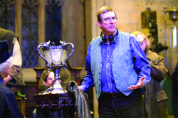 A man wearing glasses and a blue vest gestures with his hands in a room with a large silver trophy.