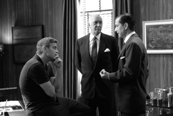 Three men in a wood-paneled room; one sits in a dark shirt while two stand in suits.