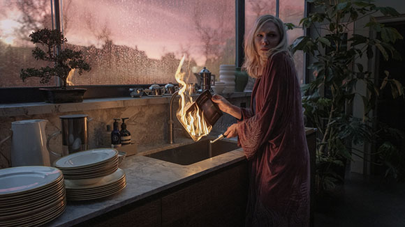 A woman in a maroon robe stands by a kitchen sink next to a small flame rising from a container.