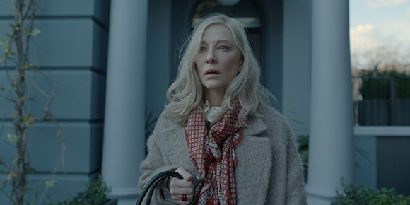 A blonde woman in a beige coat and patterned red scarf looks ahead with a surprised expression.