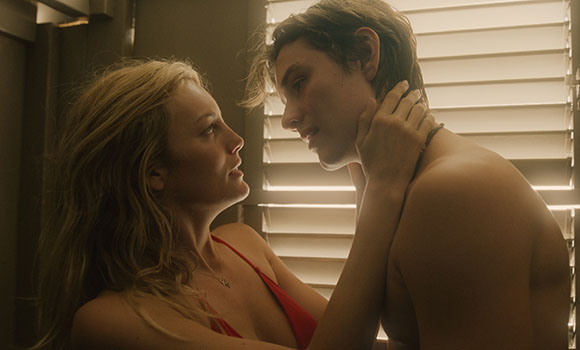 A blonde woman in a red top looks intensely at a young man as she holds his neck.