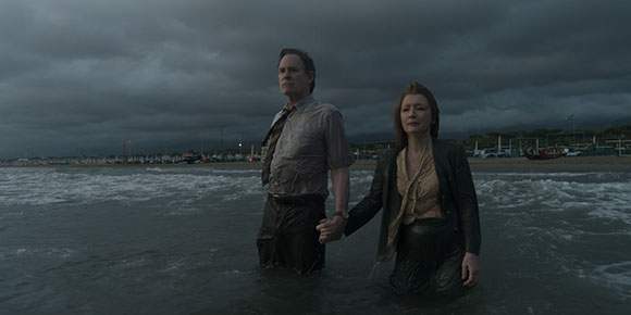 A man and woman stand waist-deep in dark ocean water under a heavy, overcast sky.