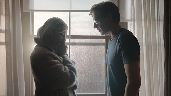 A man in a dark t-shirt looks down at a blonde woman in a heavy coat standing by a window.