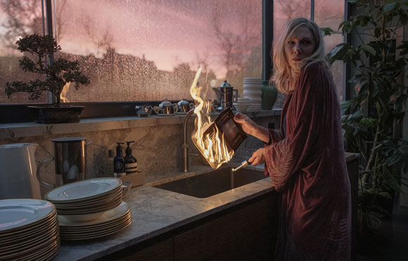 A woman in a maroon robe holds a burning book over a kitchen sink against a sunset window.
