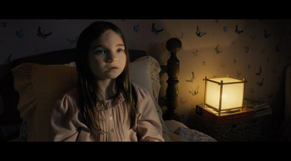 A young girl with long dark hair sits in a bed next to a glowing lamp in a dark room.