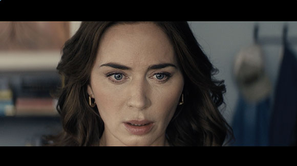 Close-up of a woman with wavy dark hair and blue eyes looking forward with a worried expression.