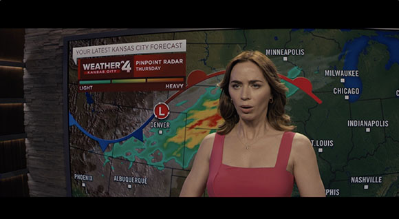 A woman in a pink top stands before a weather map showing radar patterns over the central United States.