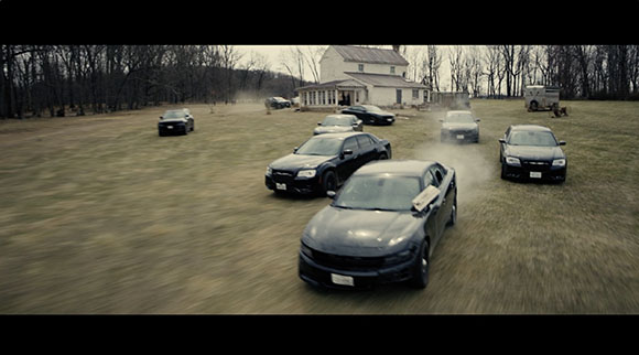 Multiple dark sedans driving across a large, grassy field toward a white house in the background.