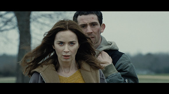 A woman with long brown hair looks forward with a worried expression while a man stands behind her.