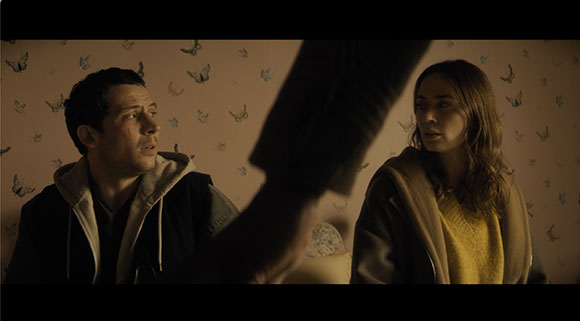 A man and a woman sit against a butterfly-patterned wall, looking toward a blurred figure in the foreground.