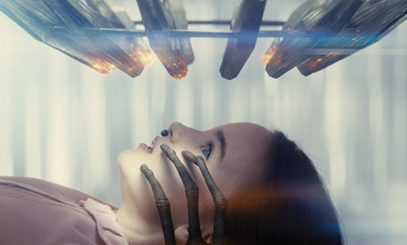A person lies on their back, looking up at large, glowing metallic structures suspended above them.