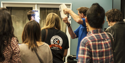 A man in a blue shirt holds up a white device while a group of people watch him. The man is demonstrating the device.