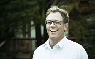 A smiling man with glasses and a light blue striped shirt standing outdoors.