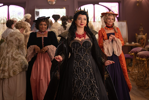 A woman in a black gown and crown stands center, flanked by two women in colorful period dresses.